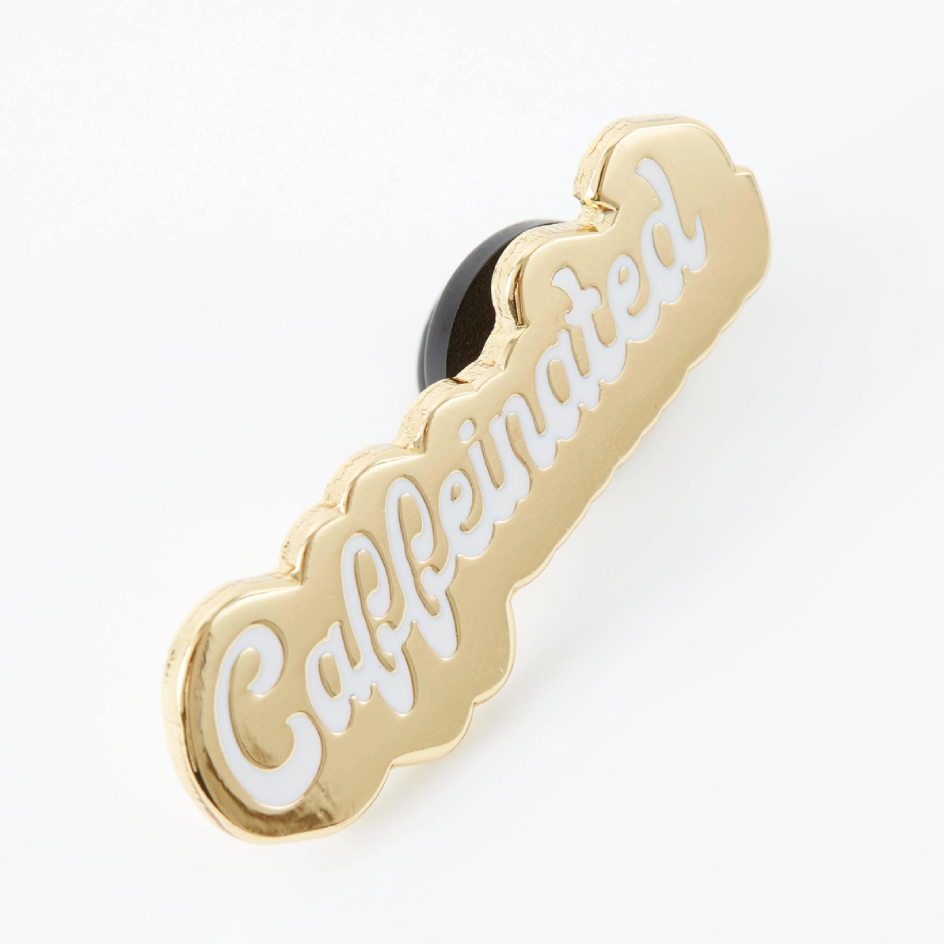 A gold colour enamel lapel pin  featuring the word 'Caffeinated' in white cursive script, shown on a clean white background.