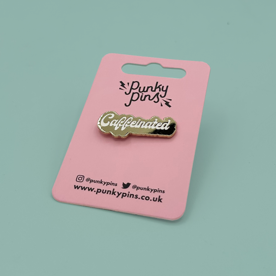 A gold-tone enamel pin with the word 'Caffeinated' in white script, mounted on a pink backing card printed with the brand name 'Punky pins'. The card is photographed on a solid light teal background.