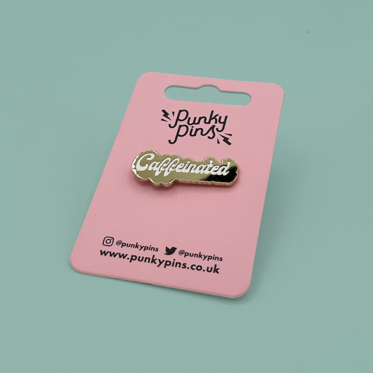 A gold-tone enamel pin with the word 'Caffeinated' in white script, mounted on a pink backing card printed with the brand name 'Punky pins'. The card is photographed on a solid light teal background.