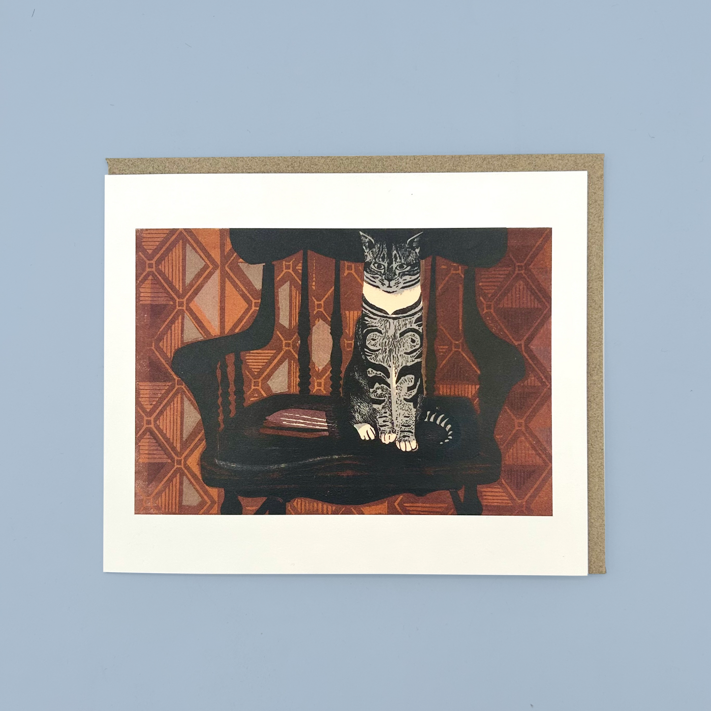 Greetings card with a linocut print of a cat sat on a chair in front of orange wallpaper. The card is on a brown envelope on a blue background.