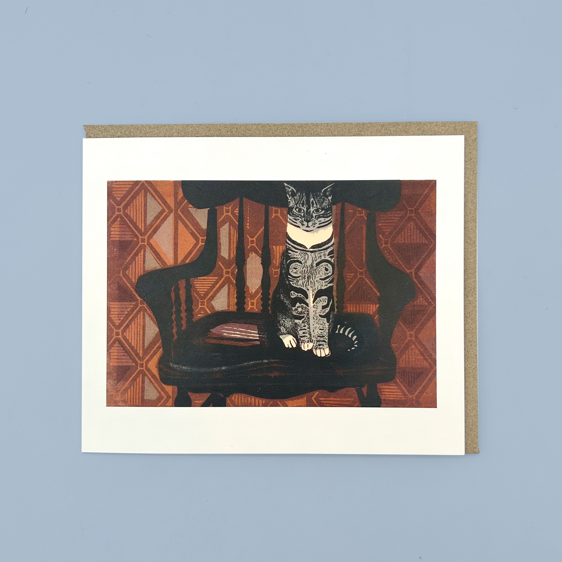 Greetings card with a linocut print of a cat sat on a chair in front of orange wallpaper. The card is on a brown envelope on a blue background.