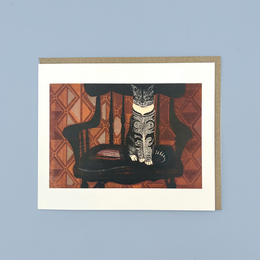 Greetings card with a linocut print of a cat sat on a chair in front of orange wallpaper. The card is on a brown envelope on a blue background.