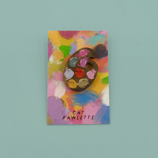 A painted wooden artist's palette pin with cat-shaped wells for paint, mounted on a colorful abstract art card labeled 'CAT PAWLETTE', set against a solid light teal background.