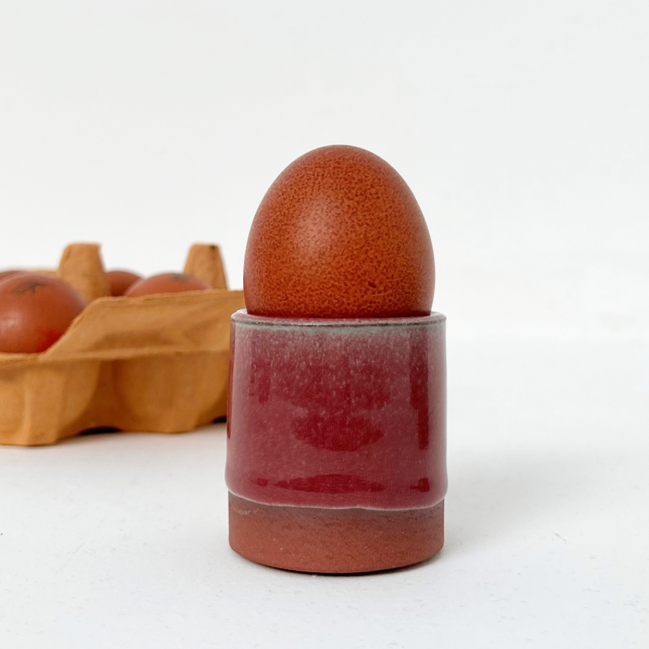 A ceramic egg holder with a glossy deep red and terracotta finish, holding a brown egg, with a blurred carton of eggs in the background on a white surface.