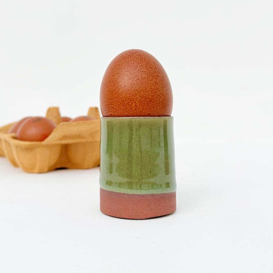 A ceramic egg holder with a glossy green middle section and terracotta-colored base, holding a brown speckled egg, with a blurred carton of eggs in the background on a white studio surface.
