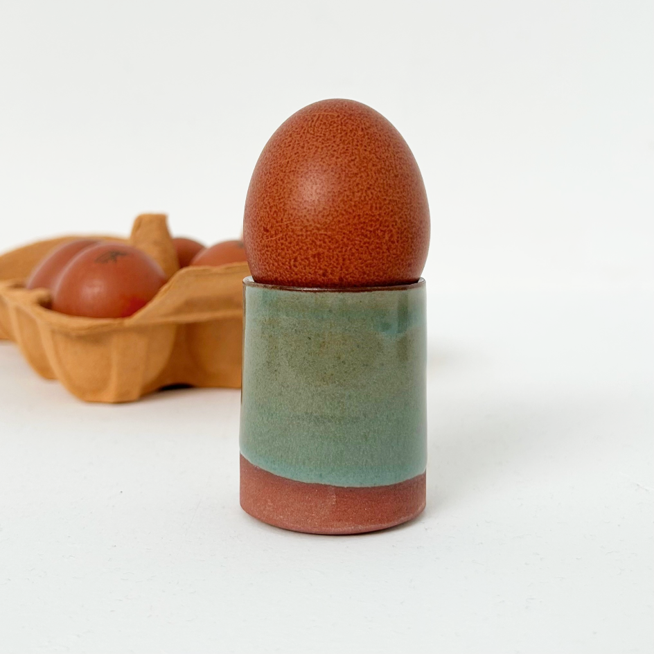 A ceramic egg holder with a glossy teal middle section and terracotta-colored base, holding a brown speckled egg, with a blurred carton of eggs in the background on a white studio surface.