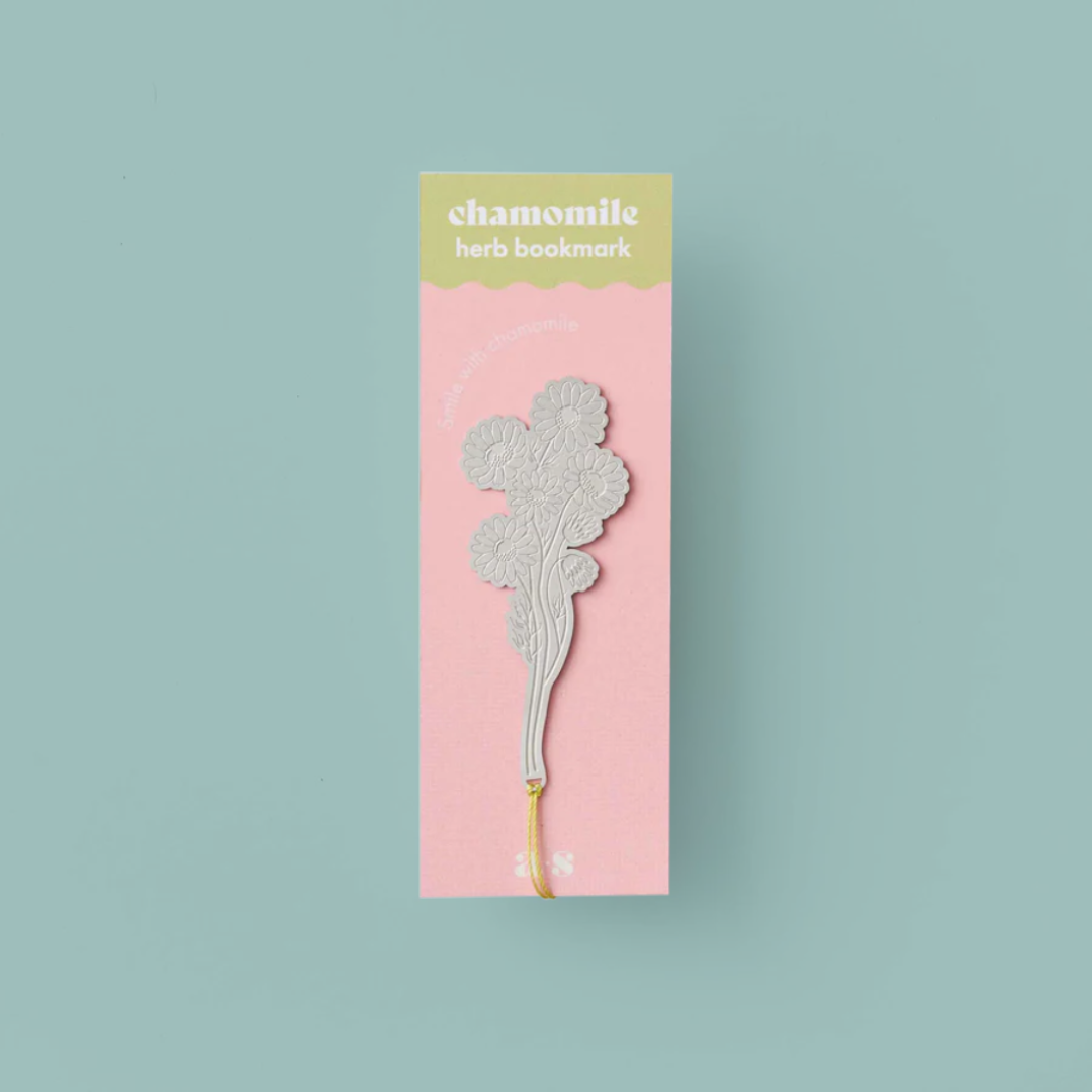 Silver coloured bookmark in the shape of a chamomile flower, in a pink and blue packet and on a light blue background. 