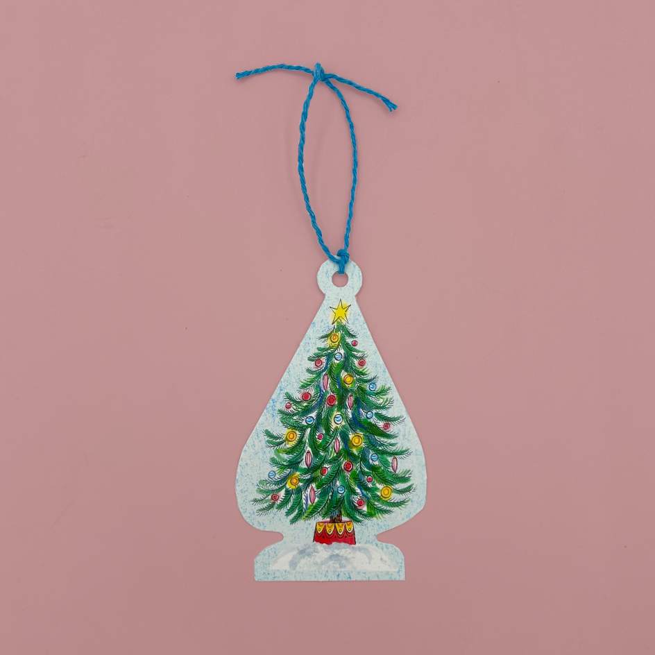 A vintage-style Christmas gift tag shaped like a teardrop, featuring a painted green Christmas tree with colorful ornaments and a red base, hanging from a blue string against a solid pink background.