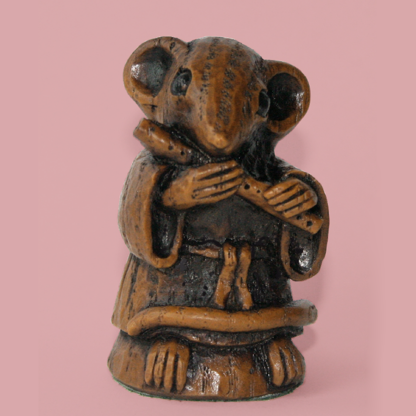 A small carved figurine of a mouse dressed in brown robes, holding a flute, set against a solid pink background.