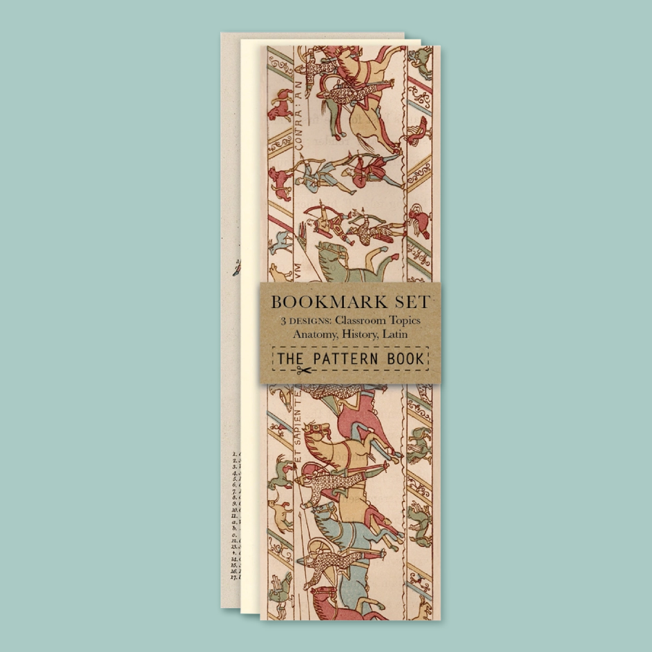 A bookmark set titled 'The Pattern Book' wrapped in a tan paper band, displayed against a solid light blue background. The bookmarks feature an illustration from the Bayeux tapestry.