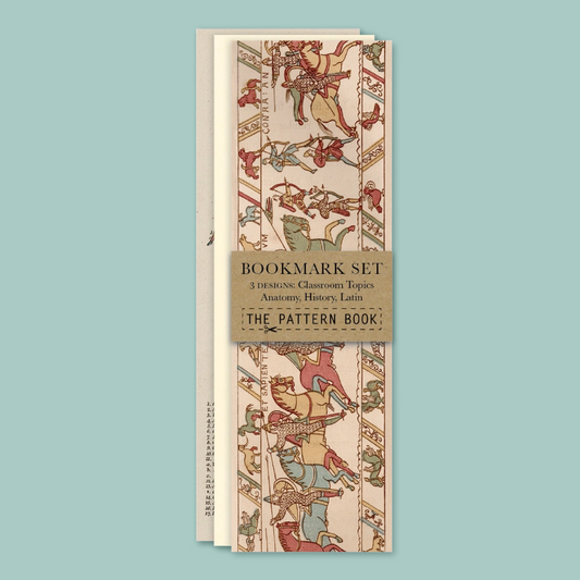 A bookmark set titled 'The Pattern Book' wrapped in a tan paper band, displayed against a solid light blue background. The bookmarks feature an illustration from the Bayeux tapestry.
