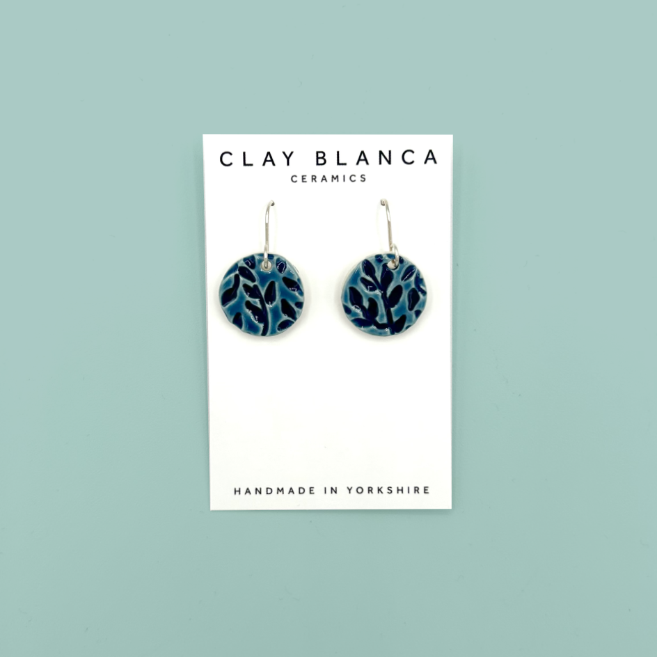 A pair of round ceramic dangle earrings with a blue leaf pattern, displayed on a white brand card that reads 'CLAY BLANCA CERAMICS'. The card is set against a solid light teal background.