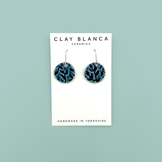 A pair of round ceramic dangle earrings with a blue leaf pattern, displayed on a white brand card that reads 'CLAY BLANCA CERAMICS'. The card is set against a solid light teal background.