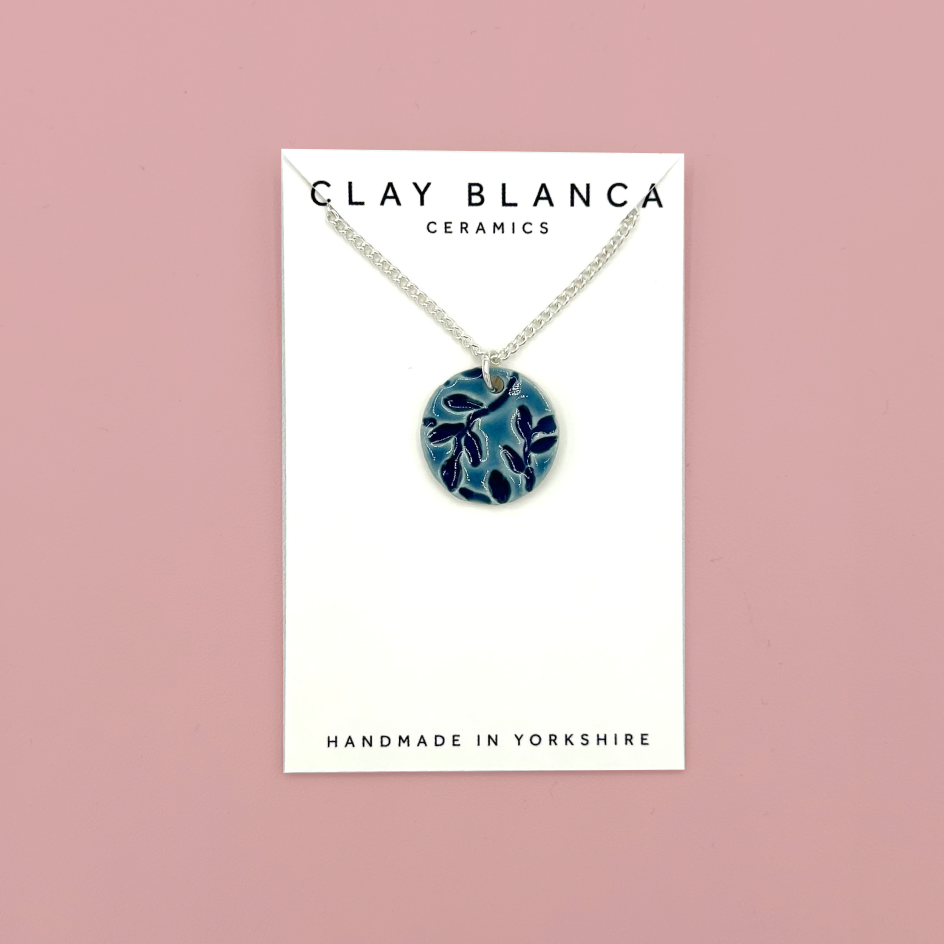 A blue patterned ceramic pendant necklace on a silver chain, displayed on a white card labeled 'CLAY BLANCA CERAMICS' against a dusty pink background.