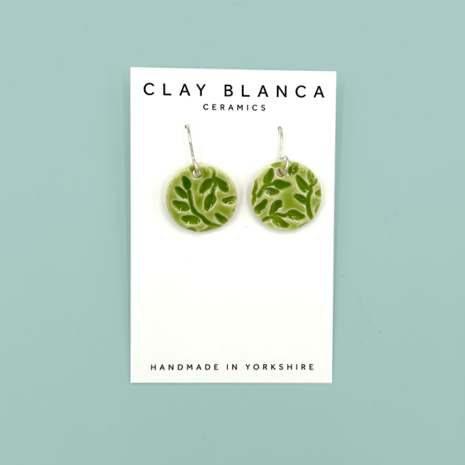 A pair of round green patterned ceramic dangle earrings on a white display card with 'CLAY BLANCA CERAMICS' printed at the top, set against a solid light teal background.