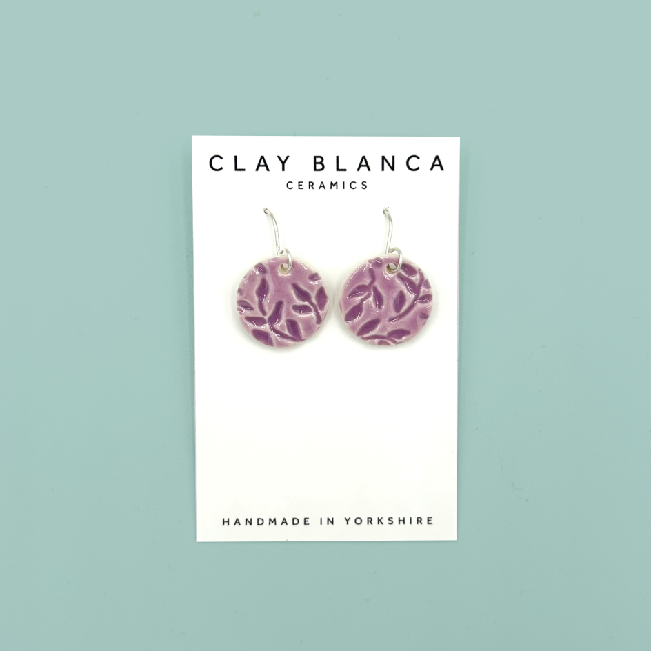 A pair of round lavender patterned ceramic dangle earrings on a white display card with 'CLAY BLANCA CERAMICS' printed at the top, set against a solid light teal background.