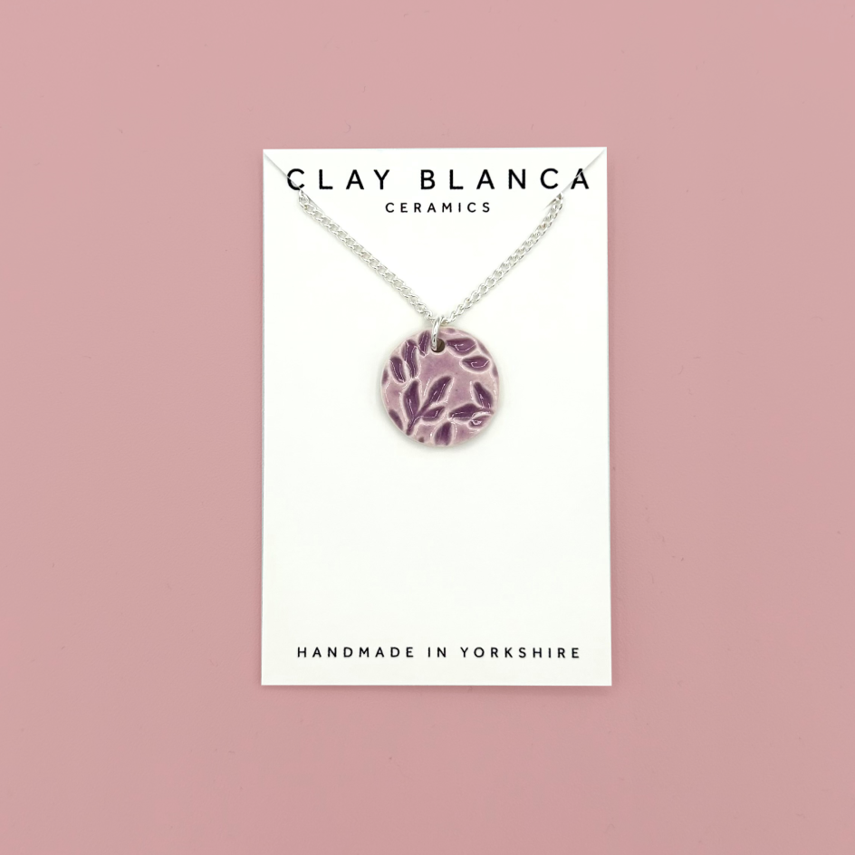 A silver chain necklace with a round pendant featuring a purple and white leaf pattern, displayed on a white card from Clay Blanca Ceramics against a dusty pink background.