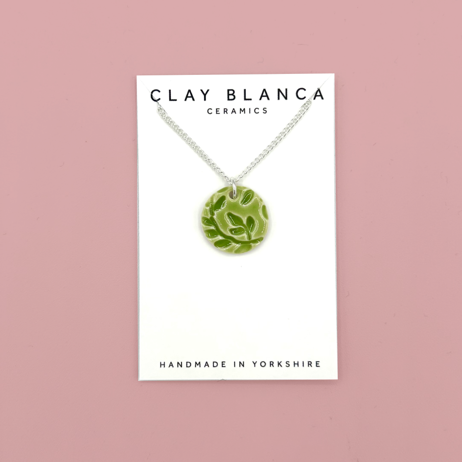 A green ceramic pendant necklace with a leaf pattern, displayed on a white card from Clay Blanca Ceramics against a dusty pink background.