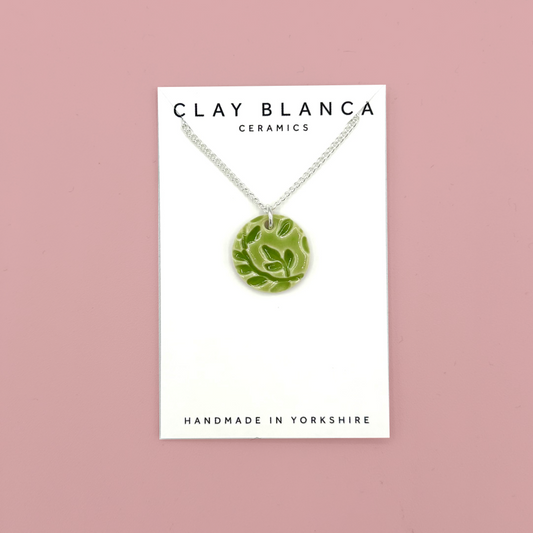 A green ceramic pendant necklace with a leaf pattern, displayed on a white card from Clay Blanca Ceramics against a dusty pink background.