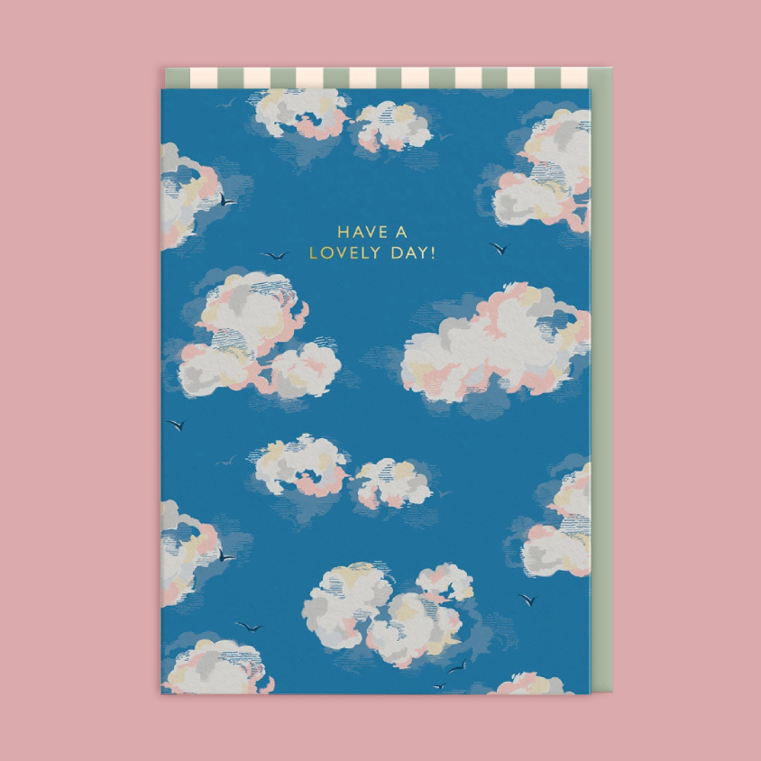 A greeting card with a blue sky and white cloud pattern, featuring the text 'HAVE A LOVELY DAY!' in gold. The card is shown against a solid pink background.