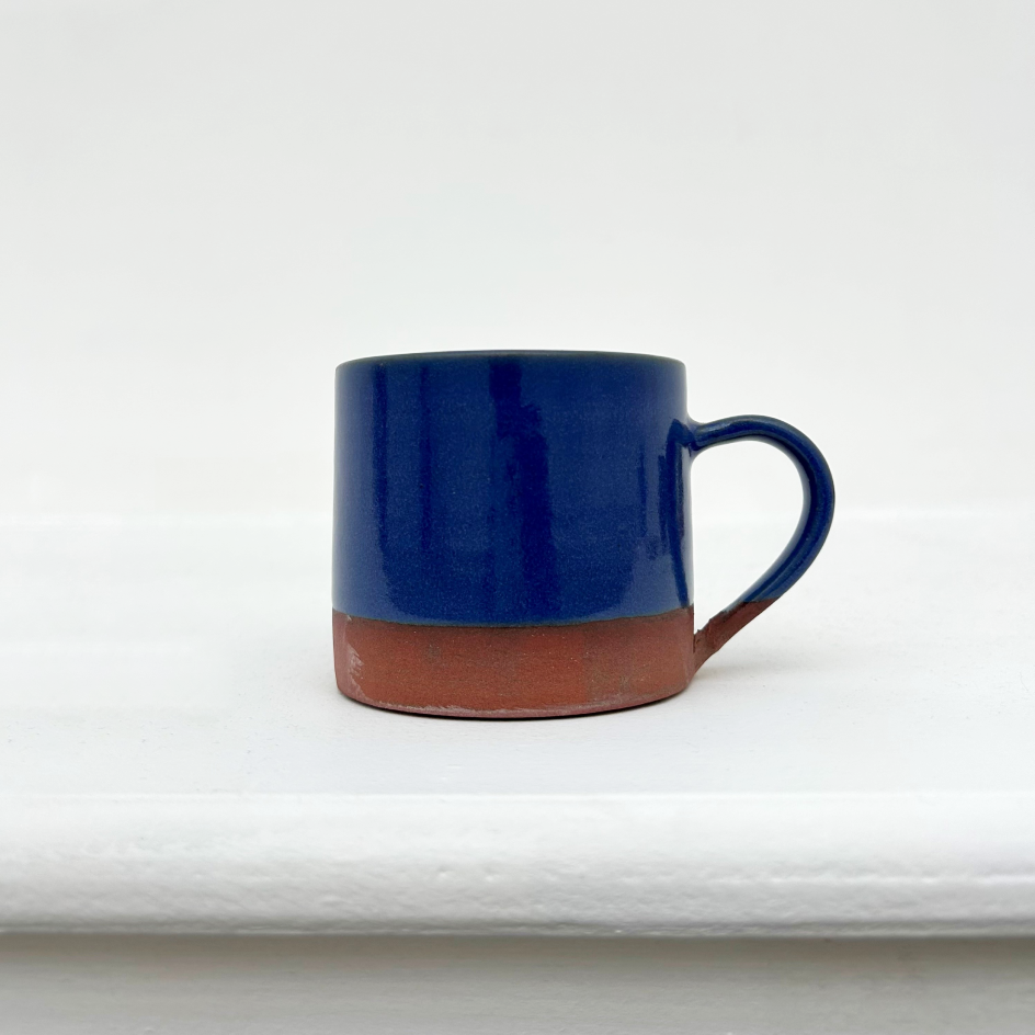 A small ceramic mug with a glossy dark blue upper body and a natural terracotta brown unglazed base, shown on a white surface against a white background.