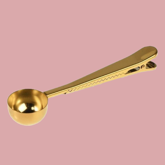 A gold-colored metal measuring spoon with a spring-loaded clip at the end, shown on a solid pink background.