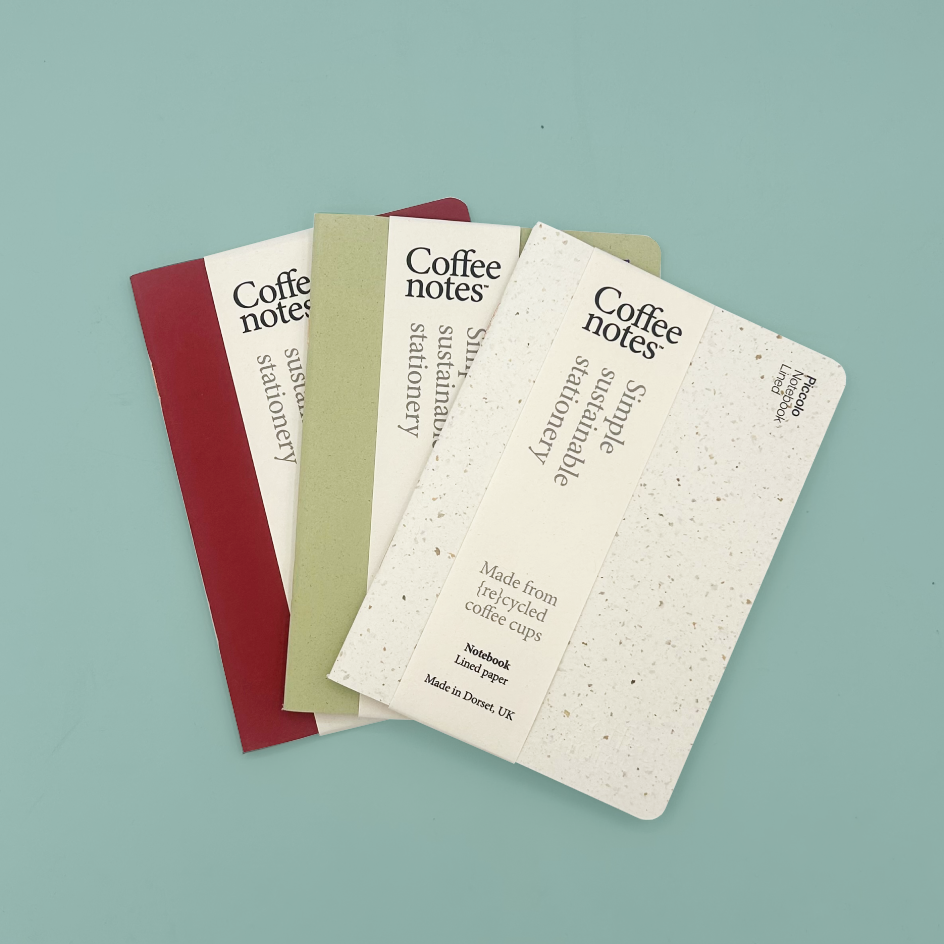 Three Coffee Notes simple sustainable stationery notebooks fanned out on a light blue background. The notebooks are made from recycled coffee cups and feature cream covers with black text, in red, green, and white/speckled paper finishes.