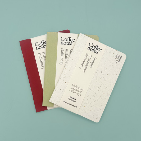 Three Coffee Notes simple sustainable stationery notebooks fanned out on a light blue background. The notebooks are made from recycled coffee cups and feature cream covers with black text, in red, green, and white/speckled paper finishes.