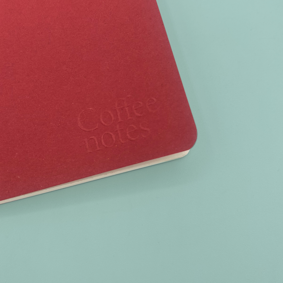 A close-up of a red notebook corner with embossed 'Coffee notes' text, placed on a light aqua background.