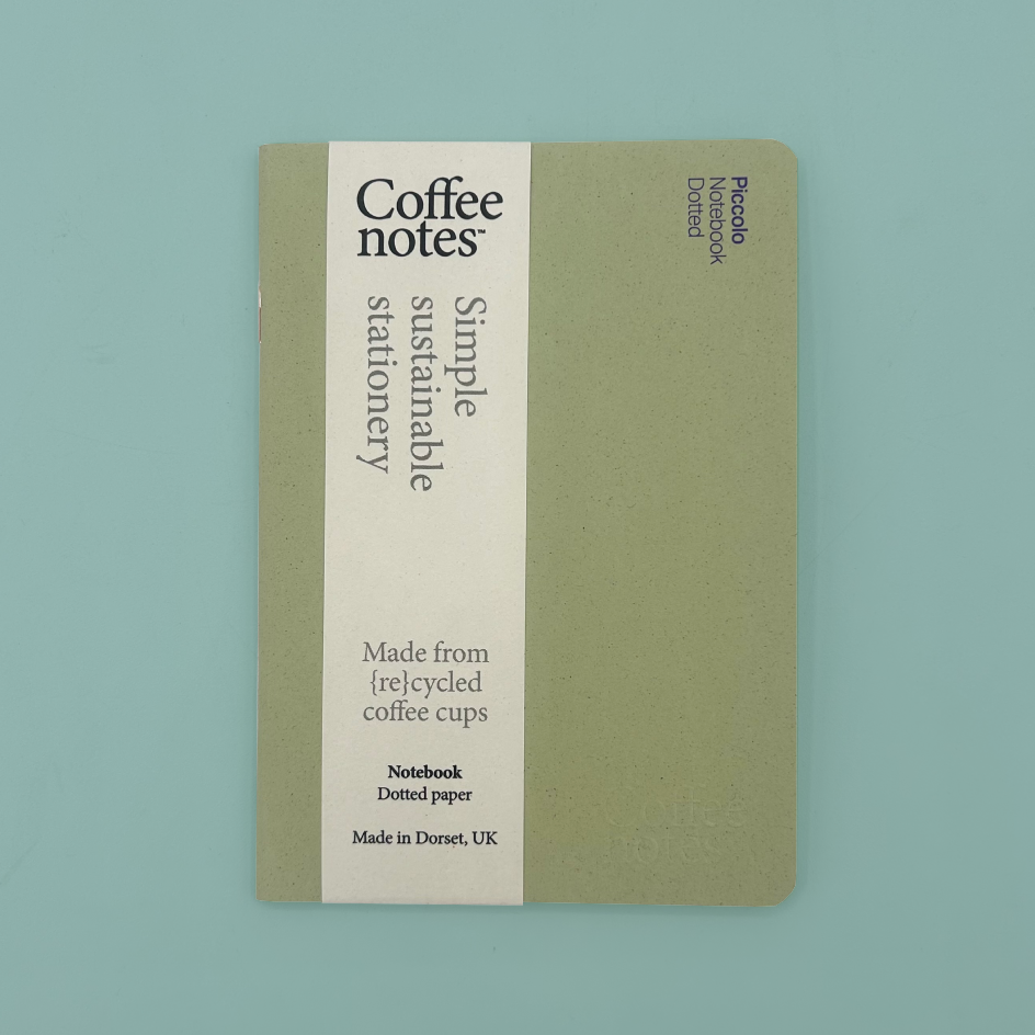 A Coffee Notes sustainable stationery notebook with a muted sage green cover and a vertical cream paper band, photographed flat on a light blue background.