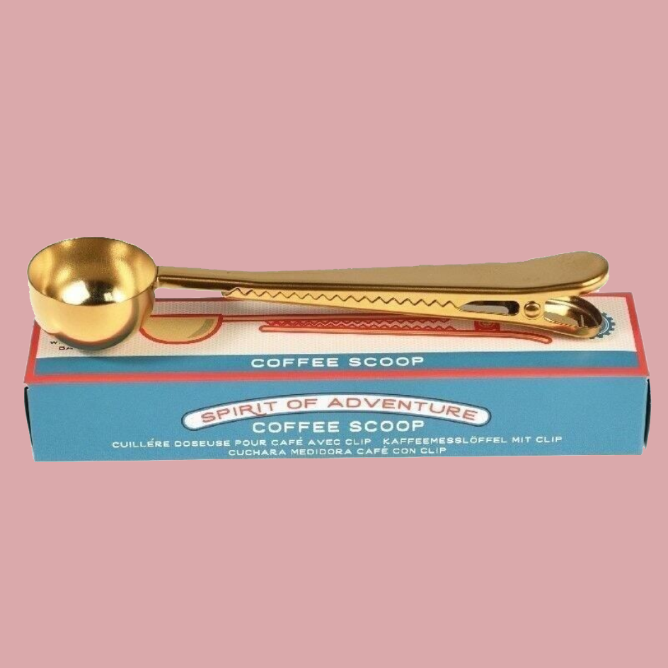 A gold-colored coffee scoop with a long handle rests on its blue and white retail box, which reads 'COFFEE SCOOP' and 'SPIRIT OF ADVENTURE'. The background is a solid, muted dusty pink.