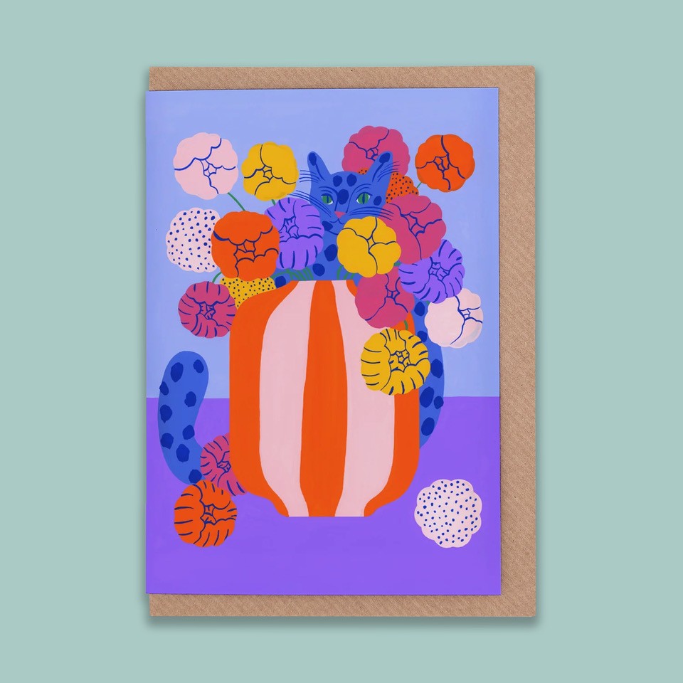 A portrait orientation greetings card with a  blue and purple background with a digital illustration of a blue spotted cat inside an orange and pink striped vase with orange, yellow, pink, and purple flowers. The card is on a brown envelope on a plain backdrop. 
