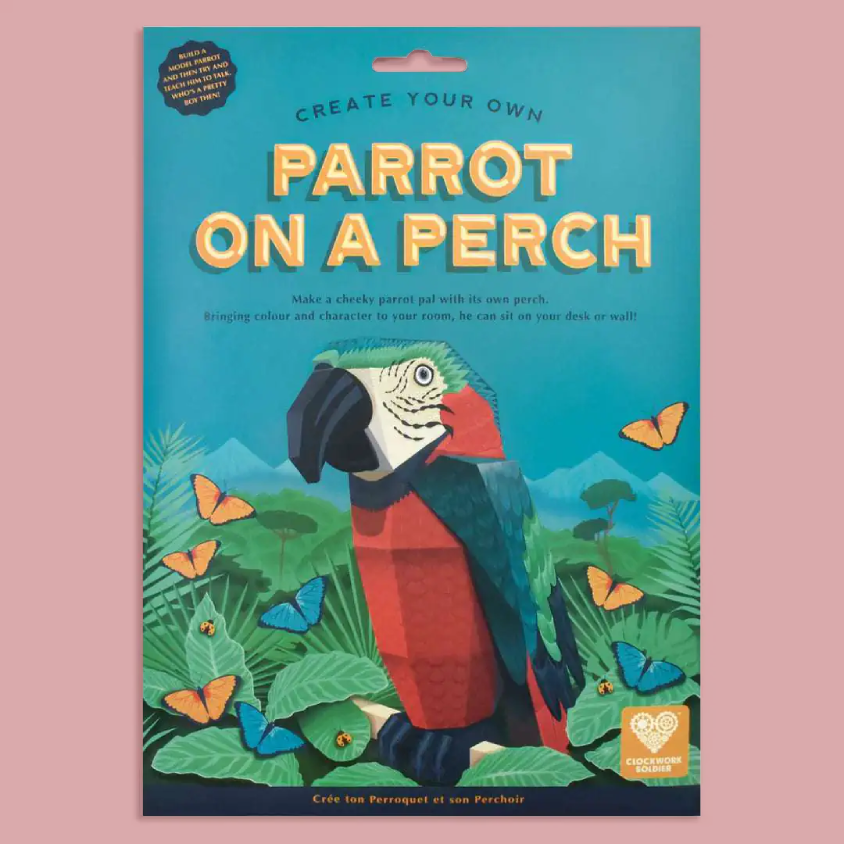 A flat-lay product shot of a craft kit package titled 'CREATE YOUR OWN PARROT ON A PERCH'. The packaging features a colorful illustration of a parrot and tropical foliage on a teal background, set against a solid pink backdrop.