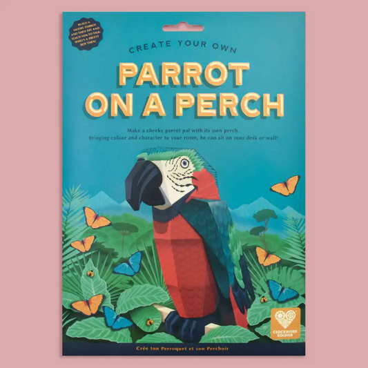 A flat-lay product shot of a craft kit package titled 'CREATE YOUR OWN PARROT ON A PERCH'. The packaging features a colorful illustration of a parrot and tropical foliage on a teal background, set against a solid pink backdrop.