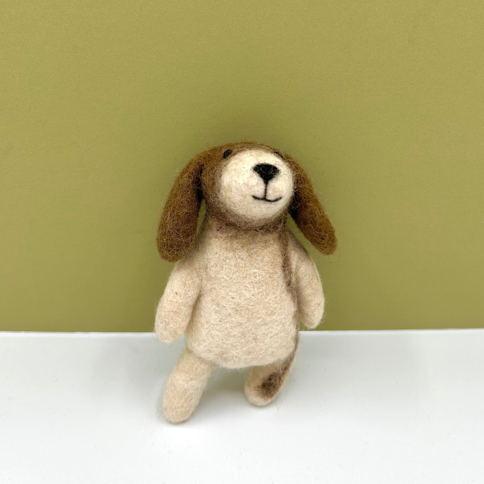 A small felted wool dog toy with long floppy brown ears and a cream-colored body, standing on a white surface against a solid olive green background.