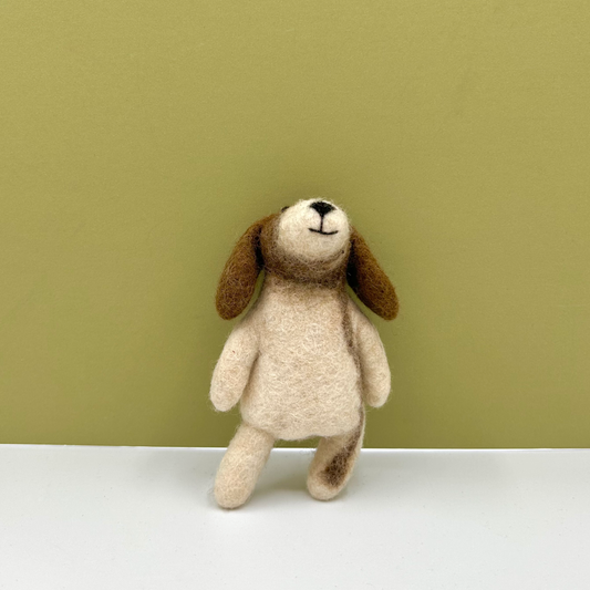A small felted wool dog toy with a cream-colored body and brown floppy ears, standing on a white surface against a solid olive green background.
