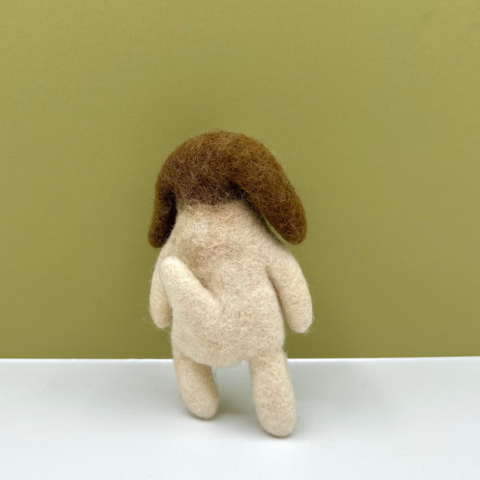 A small felted wool dog toy shown from the back, with a cream-colored body and brown floppy ears, standing on a white surface against an olive green background.