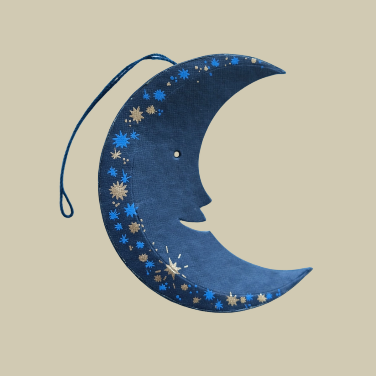 A crescent moon hanging decoration made from recycled paper, featuring a stylized face profile and blue patterned accents, suspended by a blue string against a solid light blue background.