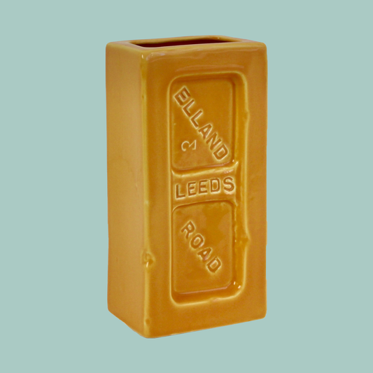 A yellow ceramic brick-shaped vase with 'Leeds Elland Road' embossed on the side.