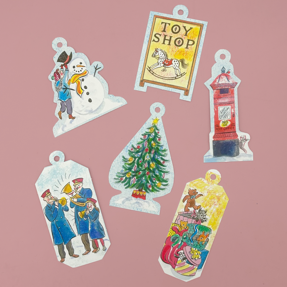Six colorful toy-themed paper cutout ornaments or tags are arranged on a dusty pink background. The designs include a snowman in snow, a toy shop sign, a red post box, a Christmas tree, and a gift box with teddy bears.