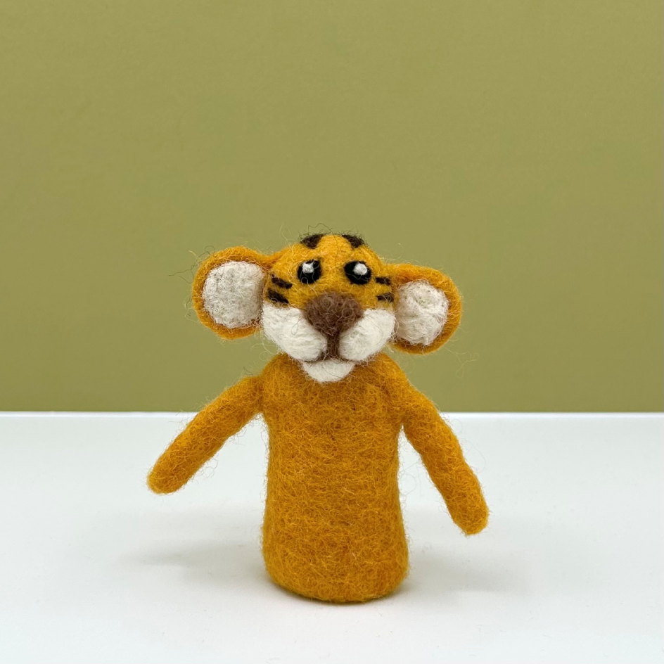 A small felted wool tiger finger puppet with orange fur, black stripes, white inner ears and face, and a brown nose, standing on a white surface against a muted olive green background.