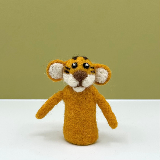A small felted wool tiger finger puppet with orange fur, black stripes, white inner ears and face, and a brown nose, standing on a white surface against a muted olive green background.