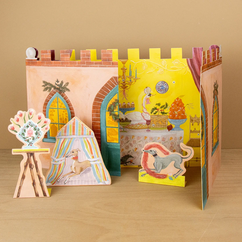 A colorful 3D pop-up advent calendar displayed on a light wood surface against a warm beige background. The set includes a castle-like backdrop with illustrated windows, a striped tent pop-up and other whimsical illustrated panels featuring animals and decorative scenes.
