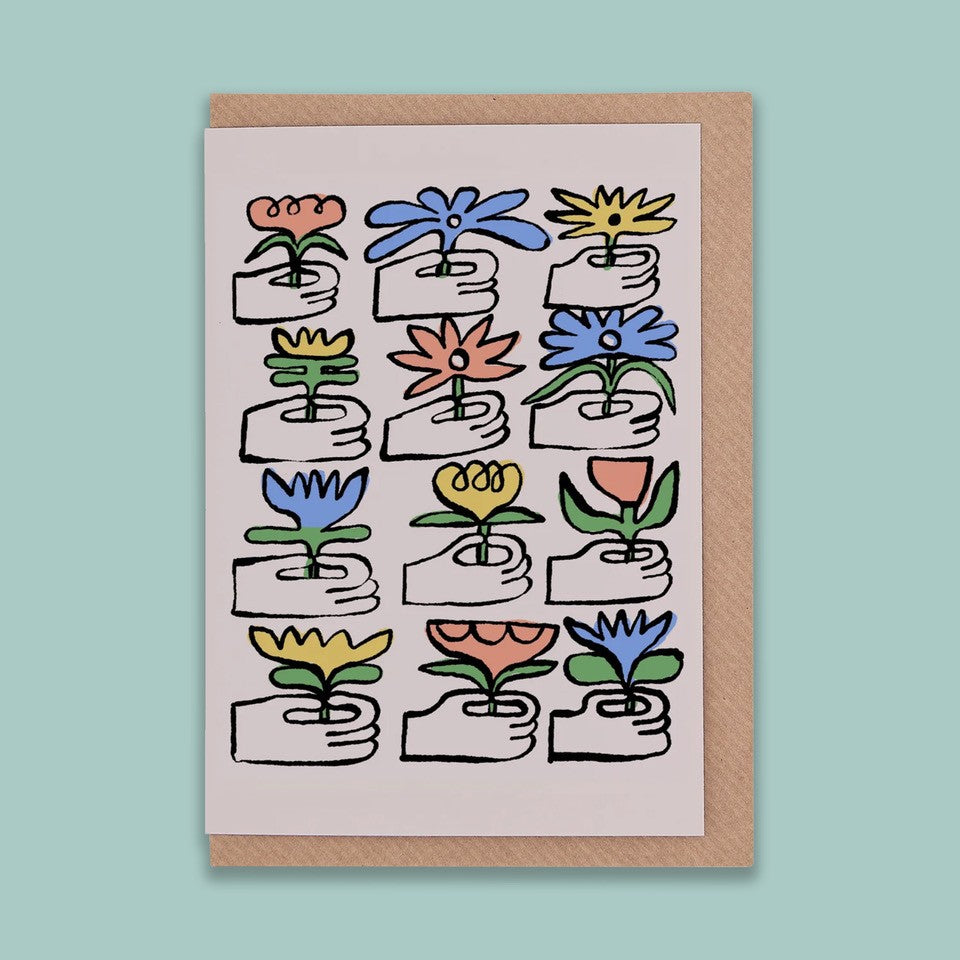 A portrait orientation greetings card with an illustration of a grid of 12 hands giving blue, red, and yellow flowers on a beige background. The card is on a brown envelope on a plain backdrop. 