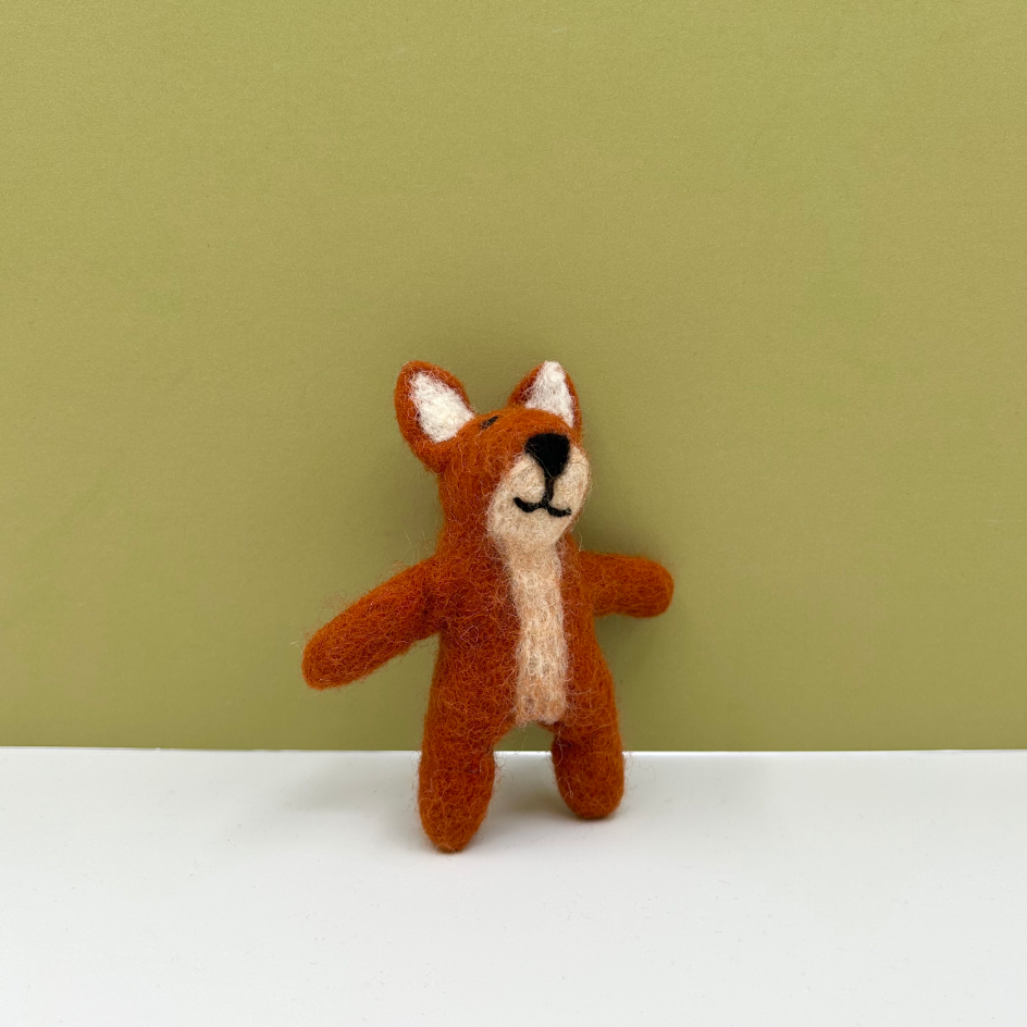 A small felted wool toy fox with orange-brown fur, cream inner ears and belly, and a black stitched face, standing on a white surface against a muted olive-green background.