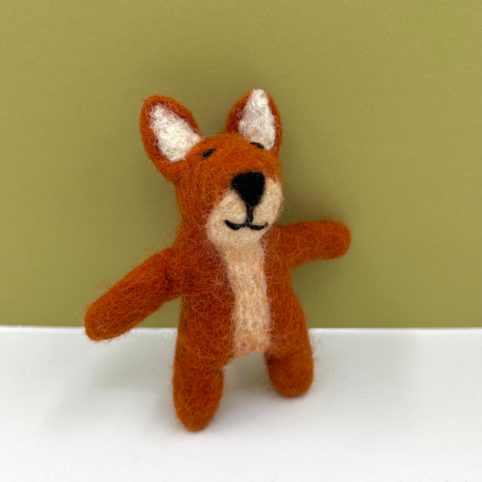 A small felted wool toy fox in rust orange with cream inner ears and a cream chest patch, standing on a white surface against an olive green background.