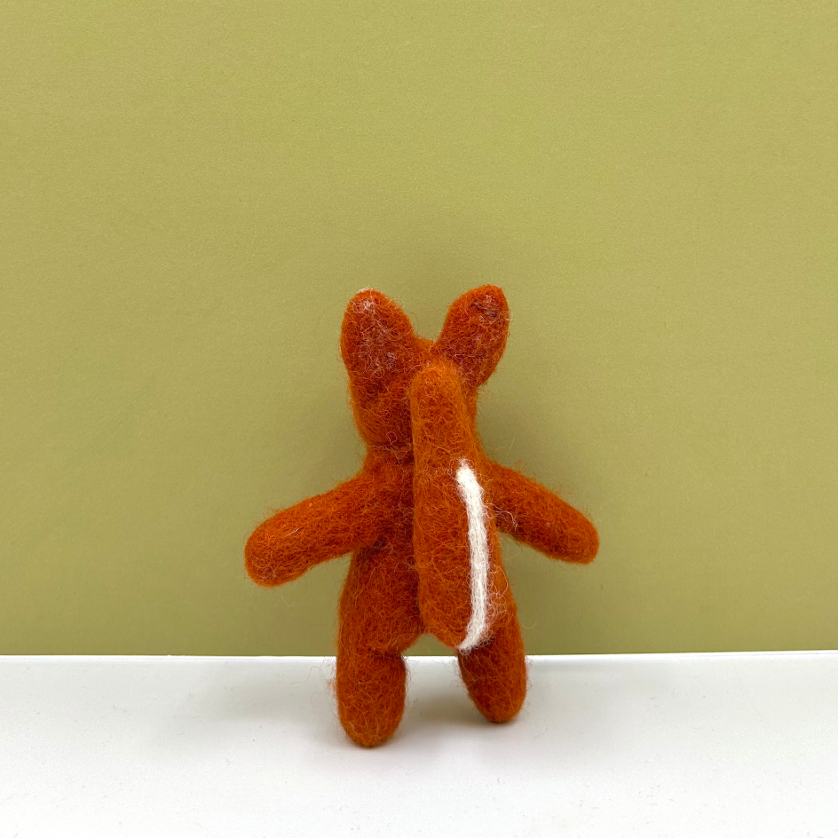 A small felted wool animal toy, shaped like a fox seen from the back, with a white stripe on its tail, standing on a white surface against a solid olive green background.