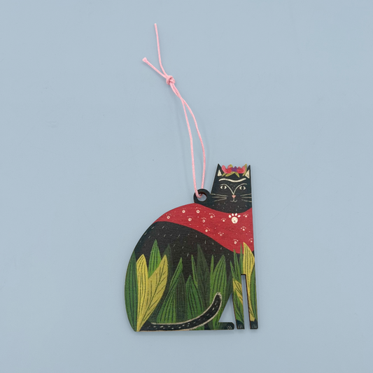 A wooden decoration shaped like a cat with black fur, green leaves, a red cape, and flowers on it's head. The decoration is hanging on a pink string and is on a blue background.
