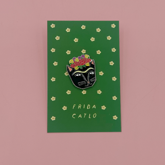 A black cat enamel pin with colorful flowers on its head, mounted on a green card with gold stars and the text 'FRIDA CATLO', set against a solid pink background.
