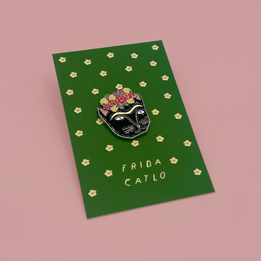 A black cat enamel pin with colorful flowers on its head, mounted on a green card with small gold flower motifs and the text 'FRIDA CATLO', set against a solid pink background.